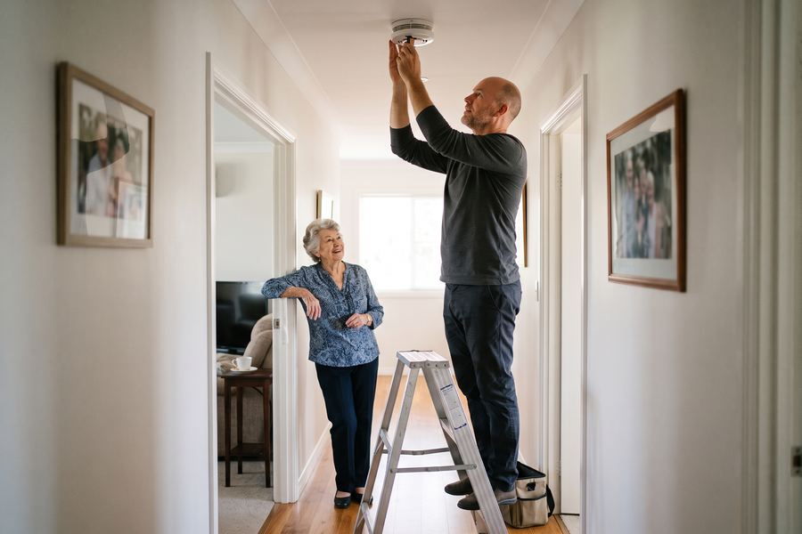 A helper on a stepladder changing a smoke alarm battery in a hallway